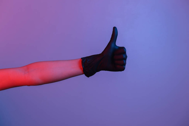 Vrouwelijke hand in zwarte latex handschoen toont duim omhoog. Gradiënt neon licht - Foto, afbeelding