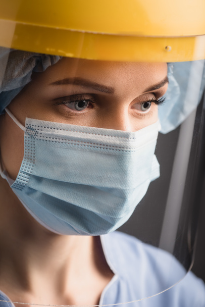 enfermera en máscara médica y protector facial aislado en gris oscuro - Foto, Imagen
