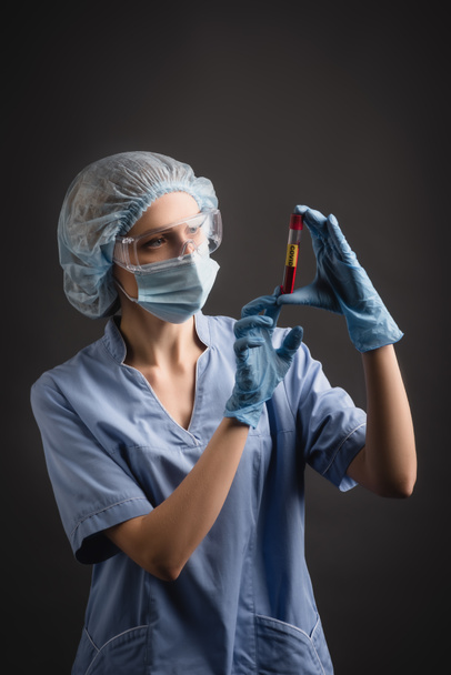 enfermera en gorra médica, máscara y gafas mirando el tubo de ensayo con letras covid aisladas en gris oscuro - Foto, Imagen