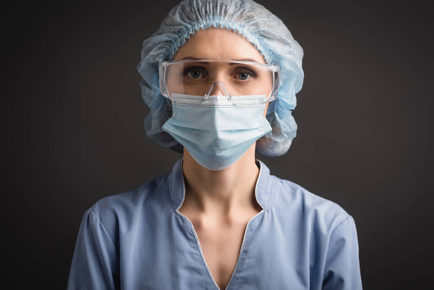 nurse in medical cap, mask and goggles looking at camera isolated on dark grey - Foto, Imagem