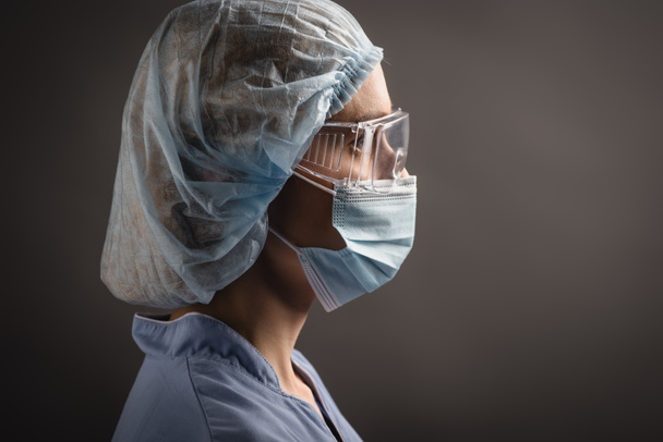 πλαϊνή όψη νοσοκόμου με ιατρικό καπέλο, μάσκα και γυαλιά που κοιτάζει μακριά και απομονώνεται σε σκούρο γκρι - Φωτογραφία, εικόνα