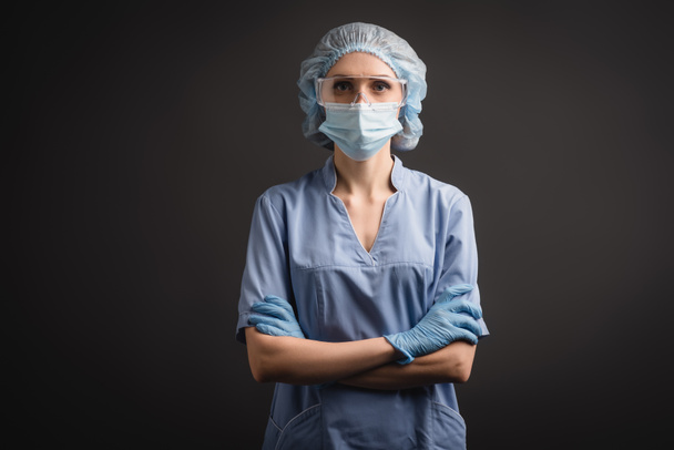 медсестра в медичній шапці, масках і окулярах стоїть з схрещеними руками і дивиться на камеру ізольовано на темно-сірому
 - Фото, зображення