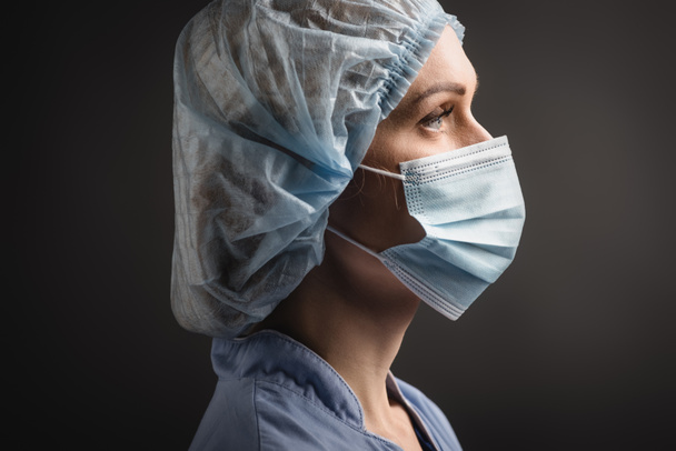 vista lateral de la enfermera en gorra médica y máscara mirando hacia otro lado aislado en gris oscuro  - Foto, Imagen