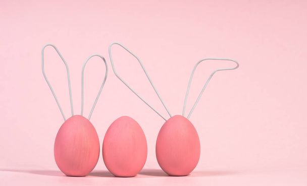 Three pink Easter eggs, cute rabbits with wire ears on a pink monochrome background with soft shadow. The concept of meeting a Happy Easter. Close-up, selective focus - 写真・画像