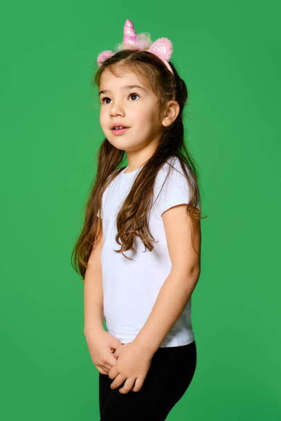 Funny little kid with unicorn hair hoop on head and in white t-shirt and black leggings posing over green background - Foto, Imagem