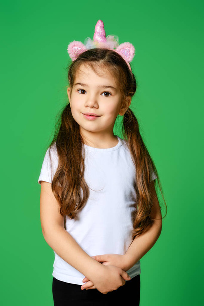 Funny little kid with unicorn hair hoop on head and in white t-shirt and black leggings posing over green background - Fotoğraf, Görsel