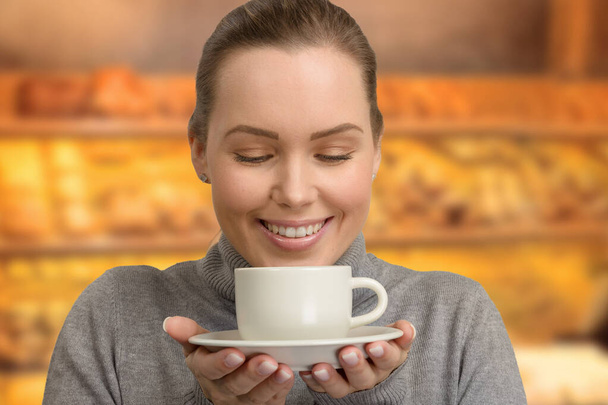 Νεαρή γυναίκα με ένα φλιτζάνι φρέσκο καφέ ή τσάι στα χέρια της μπροστά από ένα αρτοποιείο - Φωτογραφία, εικόνα