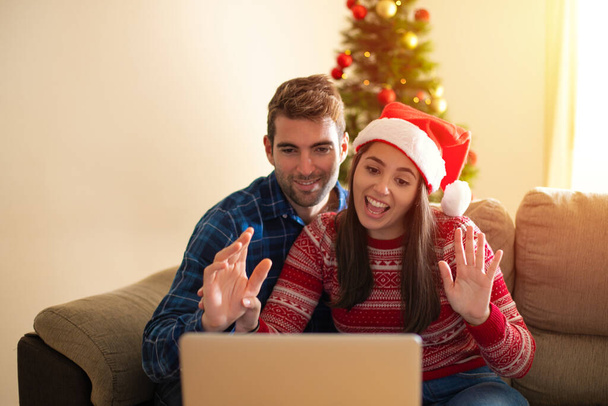 Joven hermosa pareja sonriendo feliz y confiada. Sentado en el suelo usando un portátil en una videollamada alrededor del árbol de Navidad en casa - Foto, Imagen