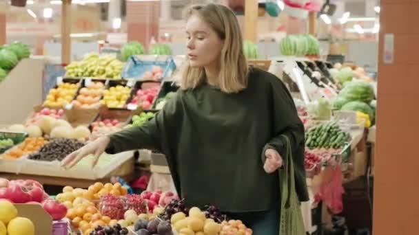 Ruční střední záběr mladé ženy se síťovou taškou výběr čerstvého ovoce a zeleniny stojí na trhu. Sbírá citron a čichá k němu. - Záběry, video