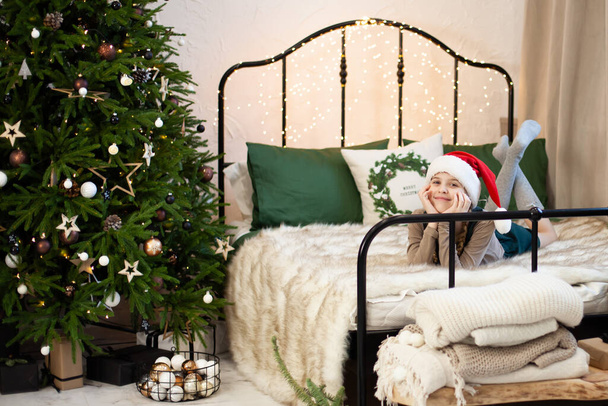 Χαμογελώντας χαριτωμένο κορίτσι στον Άγιο Βασίλη καπέλο που βρίσκεται σε ένα κρεβάτι στην εορταστική Πρωτοχρονιά και τα Χριστούγεννα ατμόσφαιρα. Μαγικό bokeh με χριστουγεννιάτικα φώτα. Καλά Χριστούγεννα και Καλή Χρονιά. Επιλεκτική εστίαση. - Φωτογραφία, εικόνα