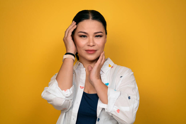 Cute mature Asian woman in a casual posing posh look on camera touching her face with hands. isolated on yellow background. Human emotions, facial expression concept - Foto, Bild