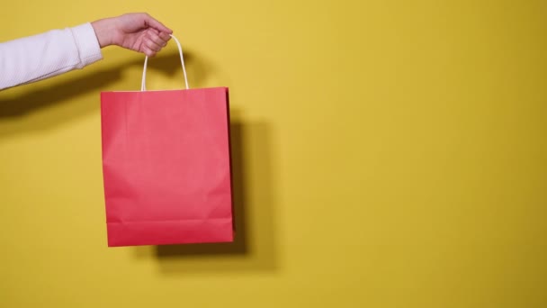 Concept d'achat ou de vente. Main féminine tient sac en papier rouge shopping isolé sur fond jaune. Bannière vidéo avec espace de copie. Prenez la main et prenez le sac en papier. - Séquence, vidéo