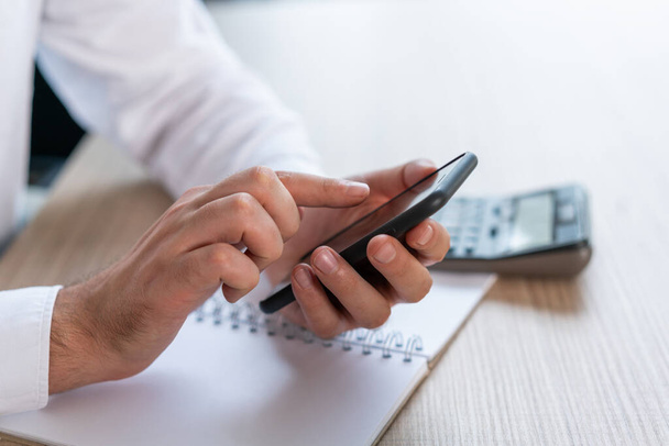 Side view of male office manager hands with black smartphone, typing a message, no face. Background of white shirt, notebook on wooden table. Concept of message - Photo, Image