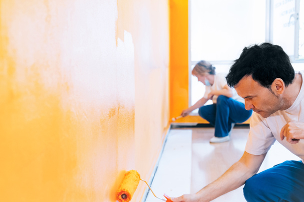 Mężczyzna i kobieta malują ścianę na pomarańczowo z wałkiem. Pracownicy malarzy wykonujący prace remontowe i reformatorskie jako zespół. - Zdjęcie, obraz