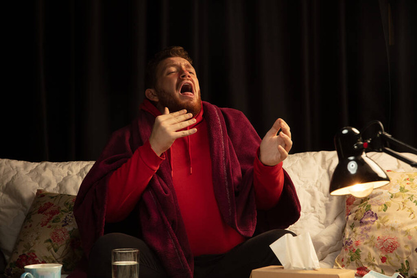 Man wrapped in a plaid looks sick, ill, sneezing and coughing sitting at home indoors - Fotografie, Obrázek
