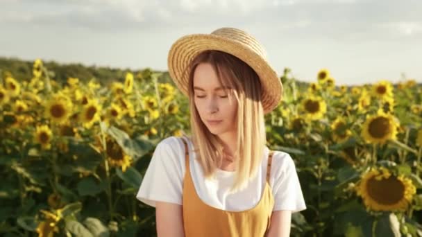 Die Frau blickt in die Kamera und lächelt. Sie trägt einen Strohhut. Sie steht inmitten von Sonnenblumen. Nahaufnahme. 4K - Filmmaterial, Video
