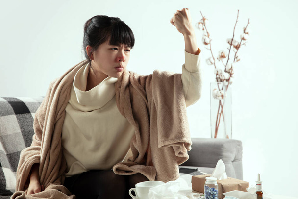 Woman wrapped in a plaid looks sick, ill, sneezing and coughing sitting at home indoors - 写真・画像