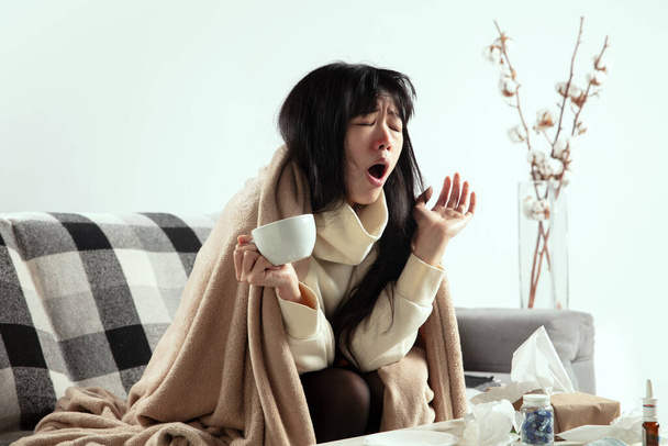 Woman wrapped in a plaid looks sick, ill, sneezing and coughing sitting at home indoors - Фото, изображение