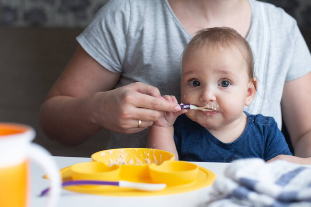 Schattige baby probeert te eten babyvoeding met behulp van lepel en zijn vrolijke moeder hem te helpen. Geel, blauw en grijs - Foto, afbeelding