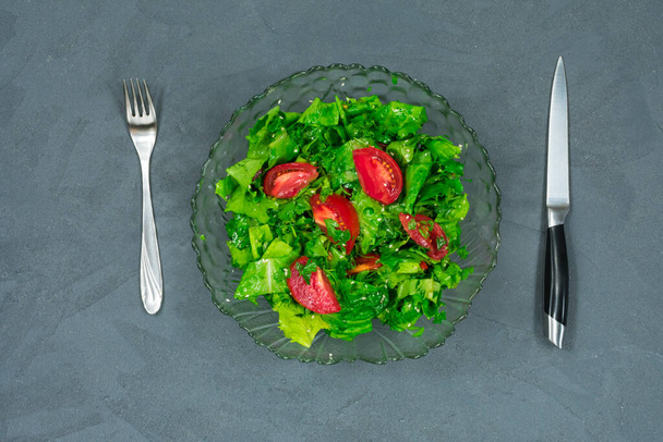 Green vegan salad made from a mixture of green leaves and tomatoes in a salad bowl with a fork and knife on a gray stone table. Top view over gray. - Photo, Image