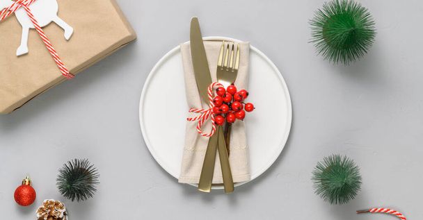 Накрытие праздничного стола с белой тарелкой, золотыми столовыми приборами и рождественскими украшениями на светло-сером столе - Фото, изображение