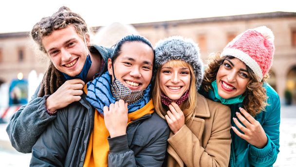Multiculturele vrienden nemen gelukkig selfie dragen gezichtsmasker en winterkleding - Nieuwe normale levensstijl concept met milenial mensen samen plezier buiten - Levendige filter met focus op blond meisje - Foto, afbeelding