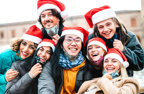 Persone multirazziali che si scattano selfie indossando maschera e cappello di Babbo Natale - Nuovo concetto di festa di Natale normale con amici felici sorridenti insieme all'esterno - Filtro retroilluminazione con focus sul ragazzo dell'Asia centrale - Foto, immagini