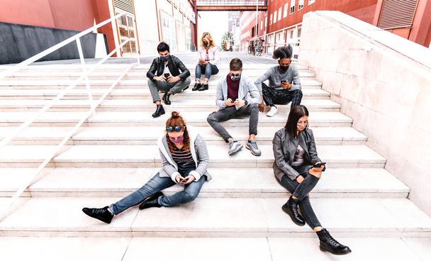 Mileniales urbanos que usan teléfonos inteligentes cubiertos por mascarilla facial en la segunda ola de Covid - Chicos y chicas preocupados viendo videos en teléfonos inteligentes - Estudiantes universitarios sentados en las vacaciones universitarias - Filtro brillante - Foto, imagen