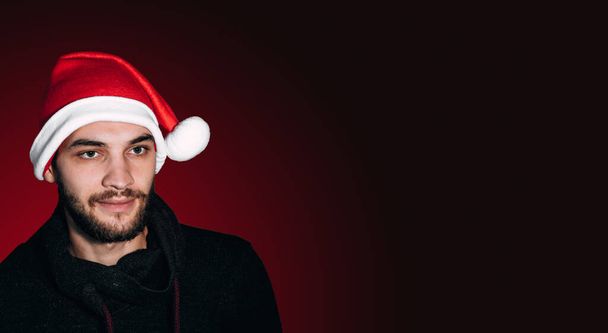 Um homem com uma barba em uma camisola de malha preta e chapéu de Papai Noel em um fundo gradiente vermelho escuro olha para o lado. Banner de Natal ano novo com um humano em um chapéu de Papai Noel vermelho. - Foto, Imagem