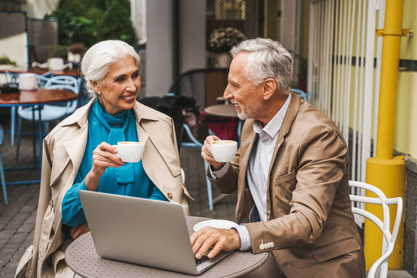Beautiful senior couple dating outdoors - Mature couple with laptop computer sitting in a bar restaurant, concepts about elderly, lifestyle and technology - Foto, imagen