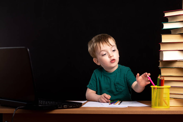 A little three-year-old boy sits at a table with books and a laptop. distance learning during quarantine. child does homework. European appearance - Photo, Image