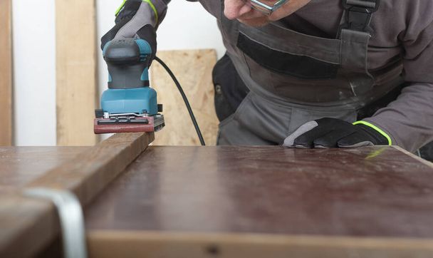 Tesař pracující s elektrickým plechem dokončovací bruska na dřevěné mříži na stole, na pozadí orientované prkno - Fotografie, Obrázek