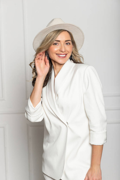 Beautiful young girl with a beautiful figure in a white trouser suit and a wide-brimmed white hat posing against a white background in the studio. woman smiling - Foto, immagini
