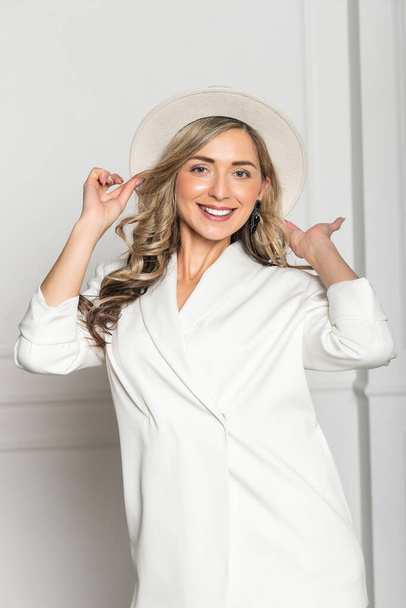 Beautiful young girl with a beautiful figure in a white trouser suit and a wide-brimmed white hat posing against a white background in the studio. woman smiling - Valokuva, kuva