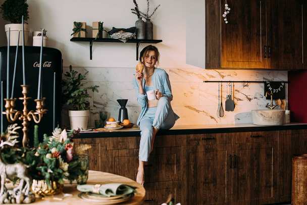 Young lady with croissant in hand chilling in the kitchen - Photo, Image