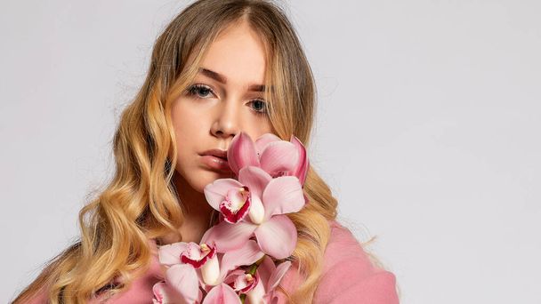 Seductive blonde woman in pink jacket posing in studio on grey background. elegant model in pastel casual spring outfit. Beautiful girl with healthy skin and branch orchid. 16:9 panoramic format. - Foto, Imagem