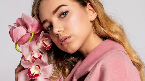 Seductive blonde woman in pink jacket posing in studio on grey background. elegant model in pastel casual spring outfit. Beautiful girl with healthy skin and branch orchid. 16:9 panoramic format. - Photo, image