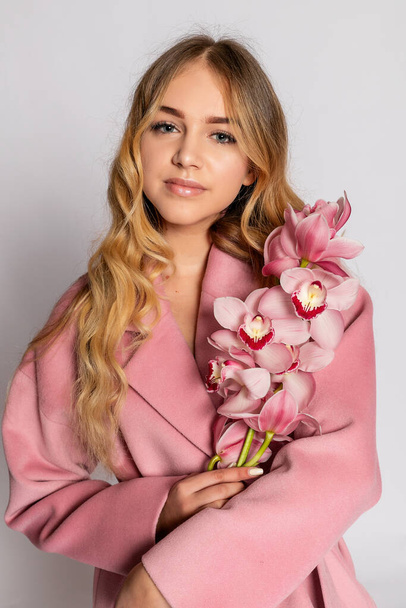 Seductive blonde woman in pink jacket posing in studio on grey background. Fashion portrait of elegant model in pastel casual spring outfit. Beautiful girl with healthy skin and branch orchid - Foto, afbeelding