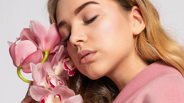 Seductive blonde woman in pink jacket posing in studio on grey background. elegant model in pastel casual spring outfit. Beautiful girl with healthy skin and branch orchid. 16:9 panoramic format. - 写真・画像