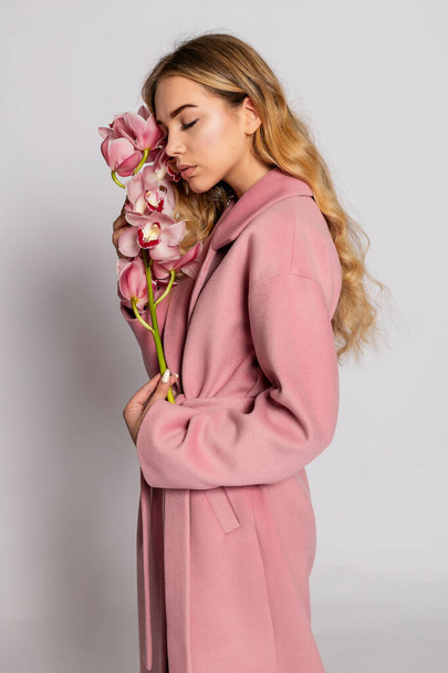Seductive blonde woman in pink jacket posing in studio on grey background. Fashion portrait of elegant model in pastel casual spring outfit. Beautiful girl with healthy skin and branch orchid - 写真・画像