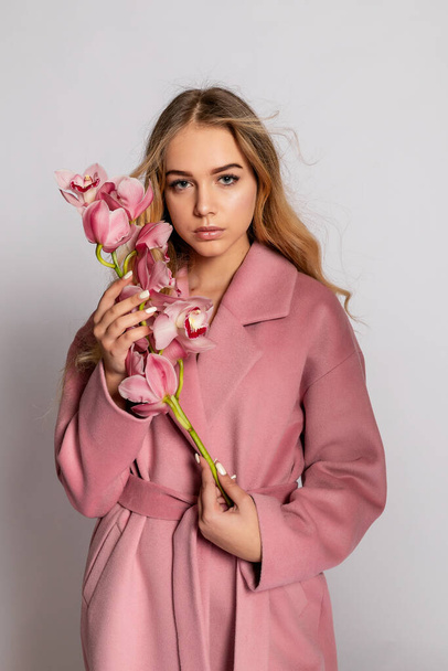 Seductive blonde woman in pink jacket posing in studio on grey background. Fashion portrait of elegant model in pastel casual spring outfit. Beautiful girl with healthy skin and branch orchid - Photo, Image