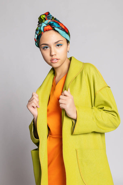Portrait of a young woman with bright makeup and a fashionable headscarf. Light background. Beauty, fashion, makeup concept. girl in a bright green coat, bright orange dress. - Fotó, kép