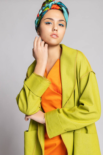 Portrait of a young woman with bright makeup and a fashionable headscarf. Light background. Beauty, fashion, makeup concept. girl in a bright green coat, bright orange dress. - Fotografie, Obrázek