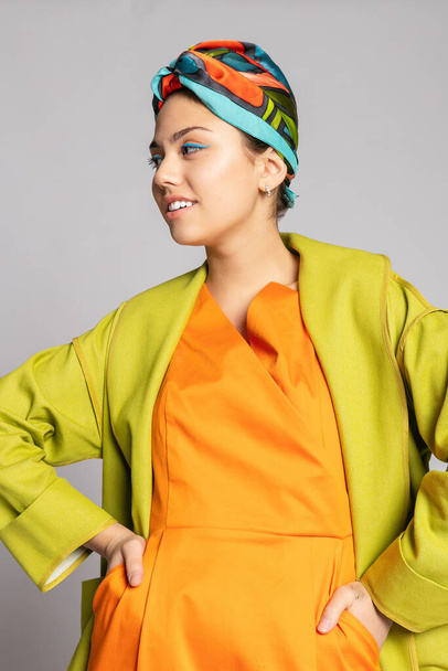 Portrait of a young woman with bright makeup and a fashionable headscarf. Light background. Beauty, fashion, makeup concept. girl in a bright green coat, bright orange dress. - Foto, Imagen