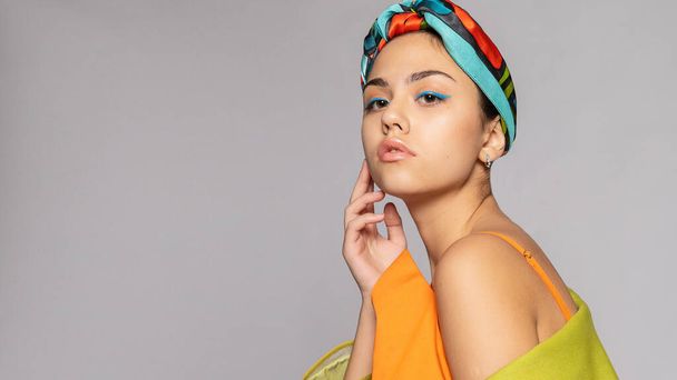 Portrait of a young woman with bright makeup and a fashionable headscarf. Light background. Beauty, fashion, makeup concept. girl in a bright green coat, bright orange dress. 16:9 panoramic format. - Fotografie, Obrázek