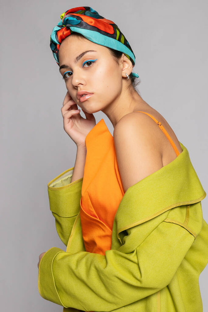 Portrait of a young woman with bright makeup and a fashionable headscarf. Light background. Beauty, fashion, makeup concept. girl in a bright green coat, bright orange dress. - Foto, Bild