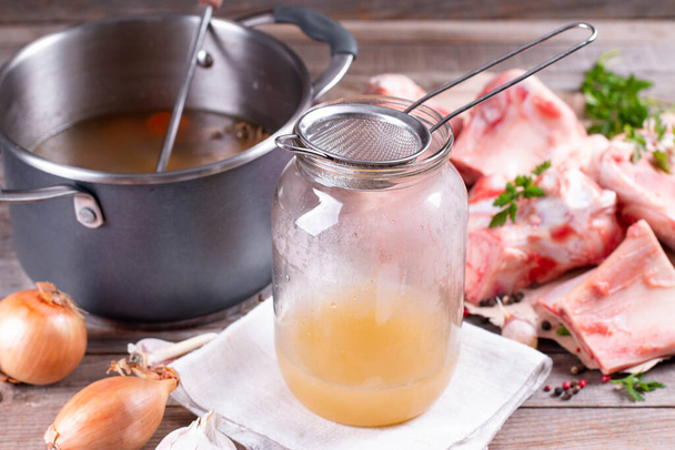 Bone Broth in a glass jar, bones and fresh vegetables, garlic cloves and herbs on wooden table, paleo diet - Photo, Image