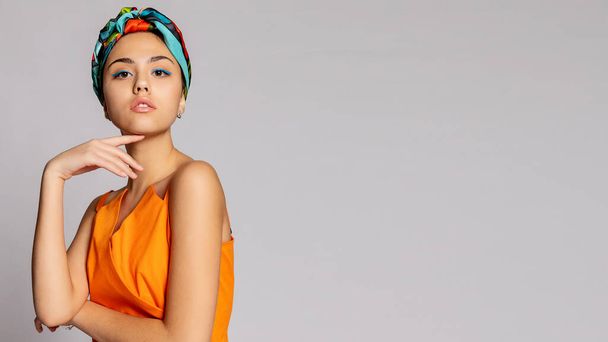 Portrait of a young woman with bright makeup and a fashionable headscarf. Light background. Beauty, fashion, makeup concept. girl in a bright green coat, bright orange dress. 16:9 panoramic format. - 写真・画像