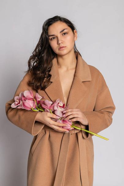 Seductive blonde woman in brown jacket posing in studio on grey background. Fashion portrait of elegant model in pastel casual spring outfit. Beautiful girl with healthy skin and branch orchid - Fotoğraf, Görsel