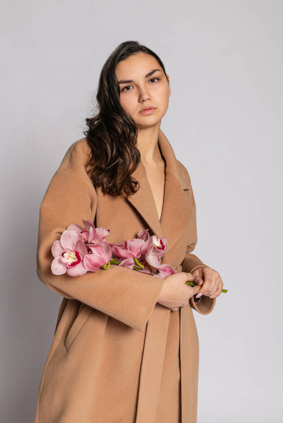 Seductive blonde woman in brown jacket posing in studio on grey background. Fashion portrait of elegant model in pastel casual spring outfit. Beautiful girl with healthy skin and branch orchid - Φωτογραφία, εικόνα